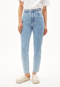 Jean taille haute bleu clair en tissu denim avec une coupe droite, doté de poches avant et d'une fermeture à bouton. Associé à des sabots beiges.