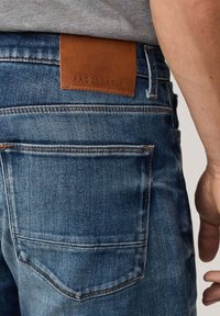 Primer plano de un bolsillo trasero de unos vaqueros azul denim con una etiqueta de marca de cuero marrón que dice "FAÇONNABLE" y una vista parcial del brazo de una persona.