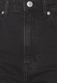 Gros plan sur un jean en denim noir montrant le bouton avant, la zone de la fermeture éclair, les passants de ceinture et les rivets argentés sur les poches.