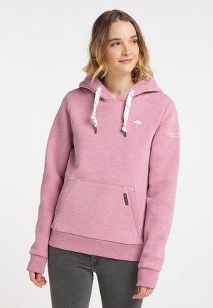 Donna che indossa una felpa rosa con cappuccio, cordini bianchi e una tasca frontale, in piedi davanti a uno sfondo chiaro e semplice.
