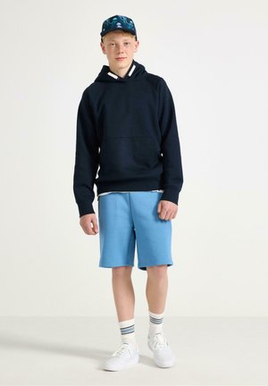 Nastolatek stoi ubrany w ciemną bluzę z kapturem, niebieskie szorty, białe trampki, paskowane skarpetki oraz niebieską baseballówkę w wzory na jednolitym tle.