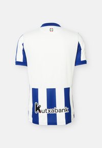 Λευκή και μπλε ριγωτή αθλητική φανέλα με κοντά μανίκια, μικρή στρογγυλή σημαία της Χώρας των Βάσκων κοντά στον γιακά και λογότυπο χορηγού Kutxabank στο κάτω μέρος της πλάτης.
