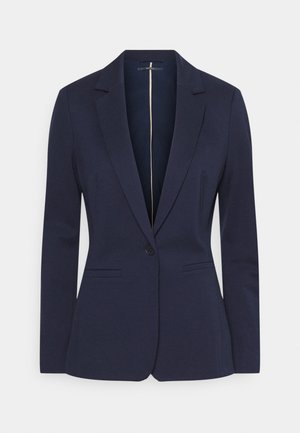 Dame blazer i navy blå med enkelt knap, notched revers og forlommer med kant, vist på ensfarvet baggrund.