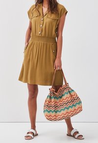 Robe à manches courtes, verte olive, avec des boutons, une taille élastique et deux poches. Grand sac à main tissé avec un motif en zigzag de plusieurs couleurs.