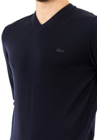 Maglione blu navy a V realizzato in morbido tessuto a maglia, con un piccolo logo Lacoste scuro sul petto e dettagli a coste sul collo.