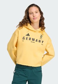 Felpa corta gialla con tasca a marsupio frontale, con scritta nera "GERMANIA", anelli olimpici e logo Adidas. Tessuto morbido, simile al cotone.