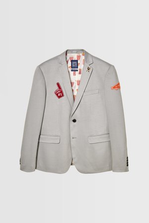 Grauer Blazer mit schwarzen Knöpfen, zwei vorderen Taschen. Enthält rot-orangefarbene Applikationen und ein gefüttertes Innenleben mit einem Popcorn-Muster.