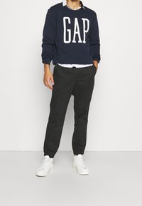 Marineblauwe katoenen sweater met "GAP" logo, zwarte tapered broek en witte sneakers. Kenmerken zijn een ronde hals en opgerolde mouwen.
