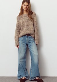 Beige neulottu pusero pitkine hihoineen, yhdistetty vaaleansinisiin leveälahkeisiin farkkuihin. Puserossa on teksturoitu pinta; farkuissa on rispaantuneet lahkeet.