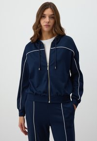 Navy blue zip-up hoodie made of soft fabric, featuring white piping along the shoulders and sleeves, with a drawstring hood and relaxed fit.
