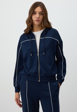Navy blue zip-up hoodie made of soft fabric, featuring white piping along the shoulders and sleeves, with a drawstring hood and relaxed fit.