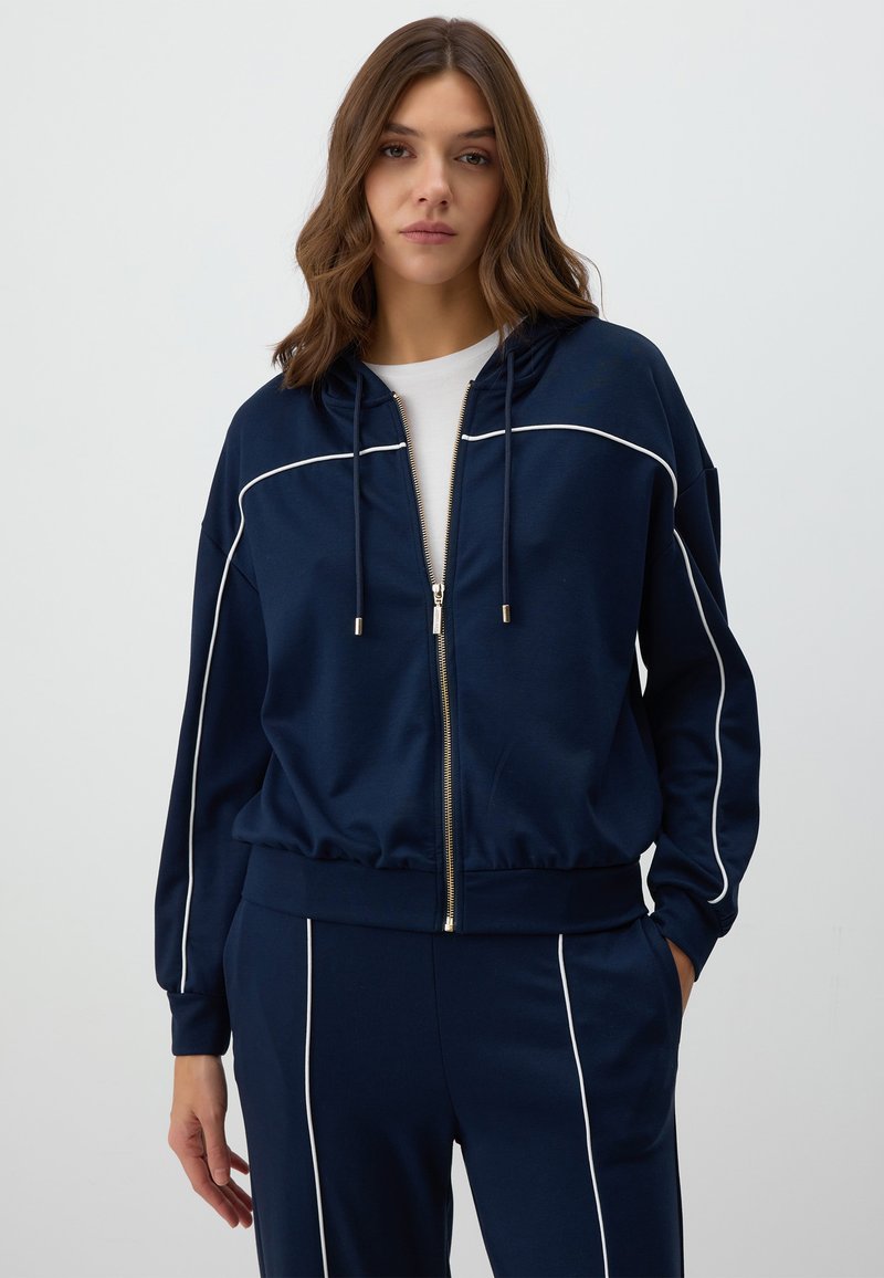 Navy blue zip-up hoodie made of soft fabric, featuring white piping along the shoulders and sleeves, with a drawstring hood and relaxed fit.