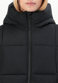 Chaqueta negra acolchada con cuello alto, que presenta un cierre central de cremallera y textura acolchada. Acabado liso sin patrones ni acentos visibles.