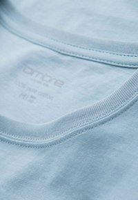 Camiseta de algodón azul claro con cuello redondo y bordes cosidos. La etiqueta indica que es 100% algodón, talla M. Textura suave.
