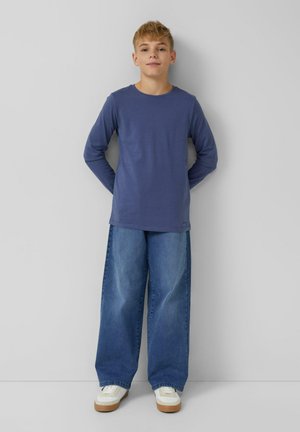 Langärmliges blaues Baumwoll-T-Shirt, kombiniert mit weit geschnittenen hellblauen Jeans. Weiße Turnschuhe mit hellbraunen Sohlen runden das Outfit ab.