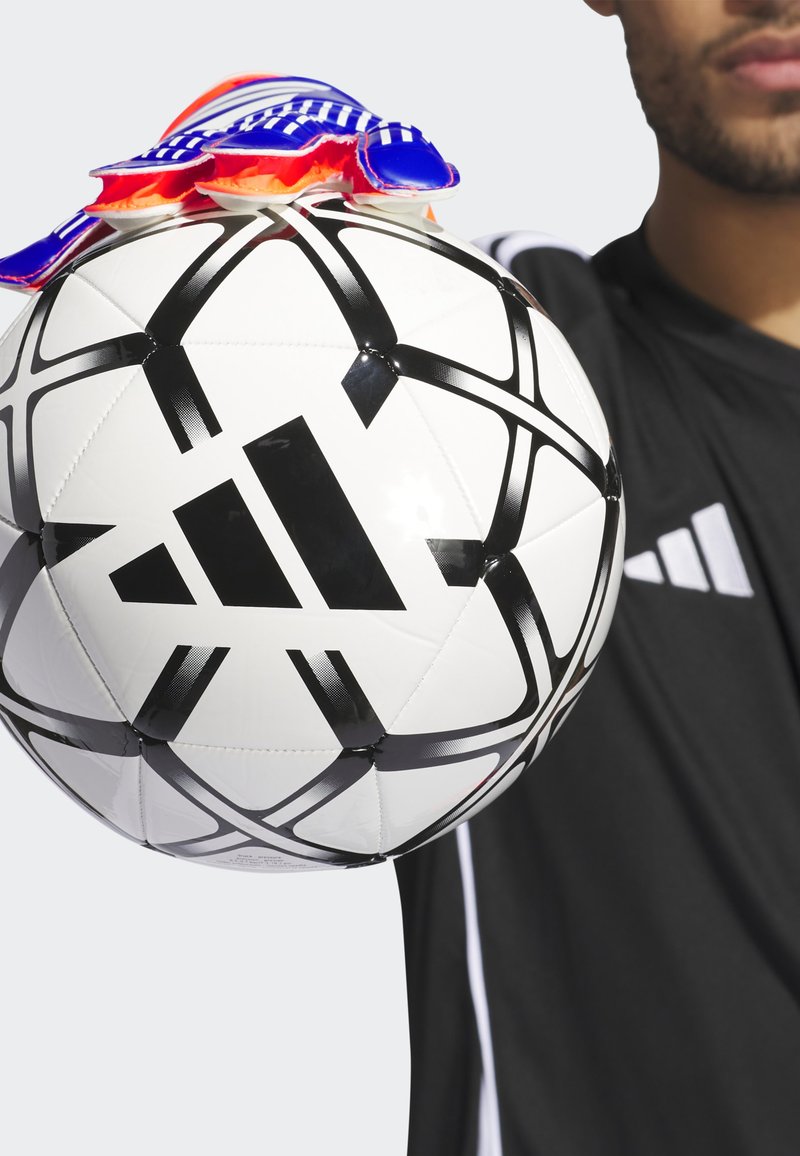 Pallone da calcio bianco con motivi geometrici neri e logo Adidas, coperto da guanti blu e rossi, tenuto da una persona in maglietta nera.