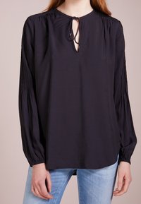Blouse manches longues bleu marine foncé avec un décolleté en forme de goutte, épaules froncées et poignets élastiques. Tissu doux avec une texture lisse.