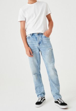 Personne portant un t-shirt blanc à manches courtes, un jean bleu clair déchiré, des baskets noires et blanches, une main dans la poche, debout devant un fond uni.