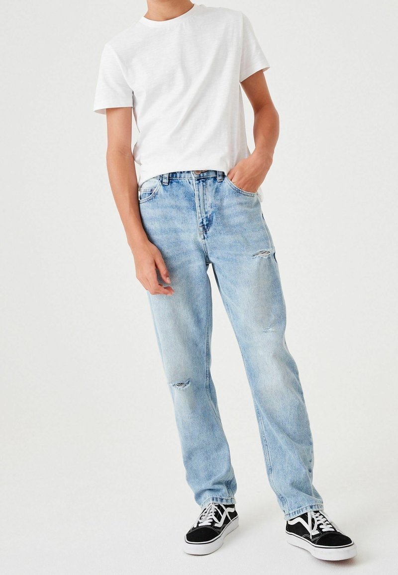 Personne portant un t-shirt blanc à manches courtes, un jean bleu clair déchiré, des baskets noires et blanches, une main dans la poche, debout devant un fond uni.