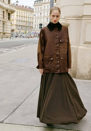 Brown padded jacket with a black collar, featuring gold buttons and pockets, paired with a long brown pleated skirt. Urban background.