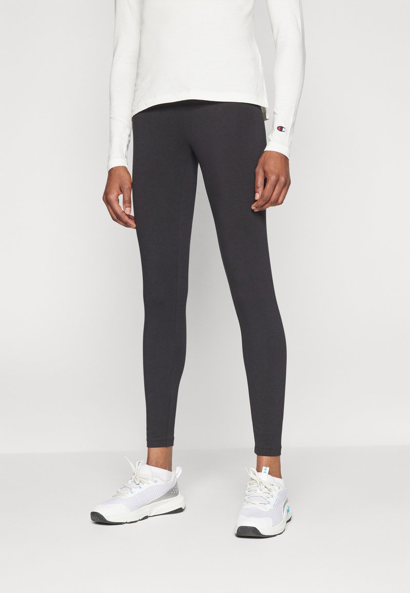 Fekete leggings elasztikus anyagból, szoros illeszkedéssel, fehér sportcipőkkel párosítva. Hosszú ujjú felső világos színben, logó részlettel.