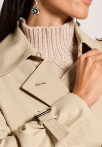 Vrouw in een beige trenchcoat over een crèmekleurige geribbelde coltrui, die de kraag van de jas vasthoudt, met zilveren bloemetjesoorbellen zichtbaar.