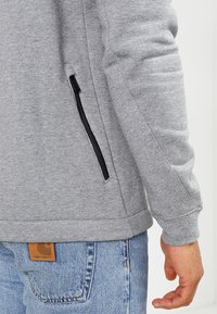 Harmaa huppari, jossa on tekstuuripinta, mustalla vetoketjullisella taskulla sivussa ja näkyväll ä bränditagilla alaselässä.