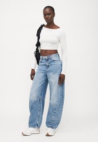 Biely dlhý rukáv crop top, svetlomodré široké džínsy s vysokým pásom a detailami šitia, nosené s bielymi teniskami. Čierna taška s volánmi.