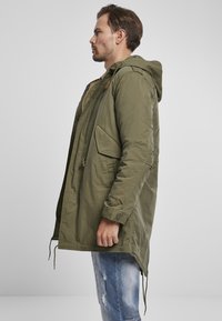 Olivově zelená parka s kapucí, disponující velkými bočními kapsami, zapínáním na knoflíky a šňůrkami na spodním lemu pro nastavitelné přizpůsobení.