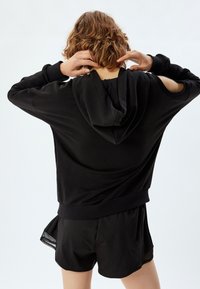 Sudadera negra con capucha de corte relajado, que presenta un diseño de mangas abiertas y un tejido texturizado. Combinada con pantalones cortos negros a juego.