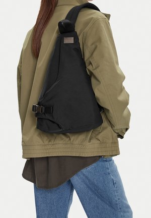 Person wearing olive jacket and blue jeans with a triangular black sling backpack over one shoulder against a white background.