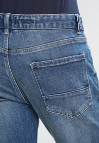 Modré džínové džíny se středním pasem, mírně vyšisovaným povrchem a zadním kapsou. Šití je viditelné kolem pasového lemu a okraje kapsy.