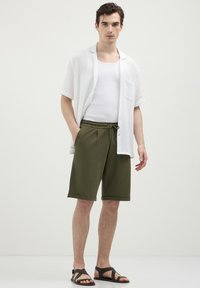 Giovane uomo in piedi con le mani in tasca, indossando una camicia bianca aperta, una canotta bianca, pantaloni corti verde oliva e sandali neri, su uno sfondo neutro.