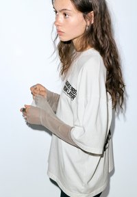 Oversized beige T-shirt med svart text, kombinerad med genomskinliga långa ärmar. Händer prydda med olika ringar, mot en vit bakgrund.