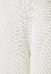 Kermanvärinen neulekangas, jossa on teksturoitu kudonta, sisältäen vinoviivat kuvioinnissa ja pehmeän tuntuman. Korosta kankaan kevyttä ulkonäköä.