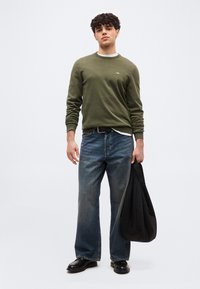 Jeune homme aux cheveux bouclés portant un pull vert olive, un jean bleu à jambes larges, des chaussures noires, tenant un grand sac noir dans la main droite.