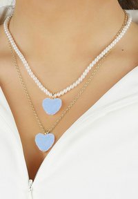 Dubbelskiktat halsband med en vit pärlsträng och en guldkedja, med två blå hjärtformade hängen, strukturerade och släta.
