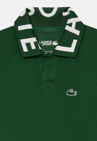 Polo shirt vert Lacoste Sport avec lettrage blanc sur le col et logo crocodile brodé sur la poitrine.