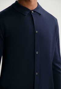 Námořnický pletený polo triko s špičatým límcem, pěti knoflíky vpředu a hladkou texturou. Jemný šev na límci.