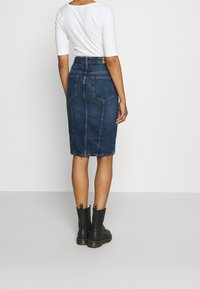 Mörkblå denimpencilkjol med hög midja, bakfickor och sömnadsdetaljer, kombinerad med svarta ankelboots. Vit åtsittande topp synlig.