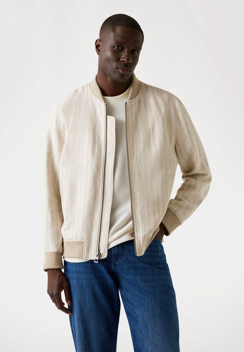 Beige bomberjack met verticale strepen, ritssluiting en ribgebreide manchetten. Gedragen over een crèmekleurig shirt en gecombineerd met blauwe spijkerbroeken.