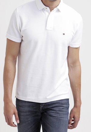 Homme portant un polo blanc à manches courtes avec un petit logo sur la poitrine et un jean bleu foncé, debout devant un fond uni.