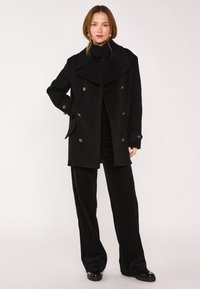 Cappotto doppio petto nero realizzato in un materiale testurizzato, con ampie patte, bottoni dorati e tasche laterali, abbinato a pantaloni neri in velluto a coste.