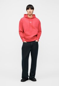 Νέος άνθρωπος στέκεται με τα χέρια στις τσέπες φορώντας κόκκινο φούτερ με κουκούλα, χαλαρό μαύρο παντελόνι και μαύρα παπούτσια μπροστά σε απλό φόντο.