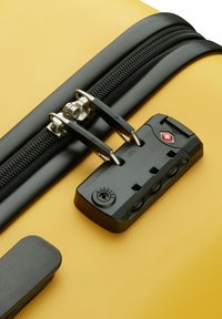 Gele bagage met een structuur oppervlak, voorzien van een zwarte rits en vergrendelmechanisme. Metalen ritsen zijn zichtbaar, met een beveiligingslabel op het slot.