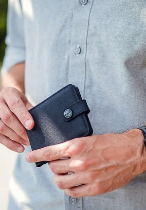 Schwarzes Portemonnaie aus strukturiertem Material, mit einem Druckknopfverschluss und einem Riemen. Wird in einer Hand gehalten und kontrastiert mit einem grauen Hemd.
