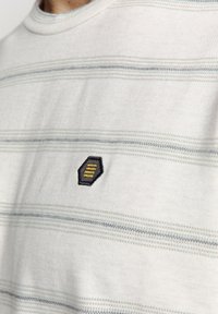 Gestreept T-shirt in het wit met blauwe en grijze horizontale lijnen, met een hexagonaal logo-embleem in zwart en geel op de linkerborst.