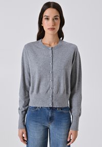 Giovane donna che indossa un cardigan grigio con bottoni e jeans blu, in piedi contro uno sfondo chiaro e neutro.