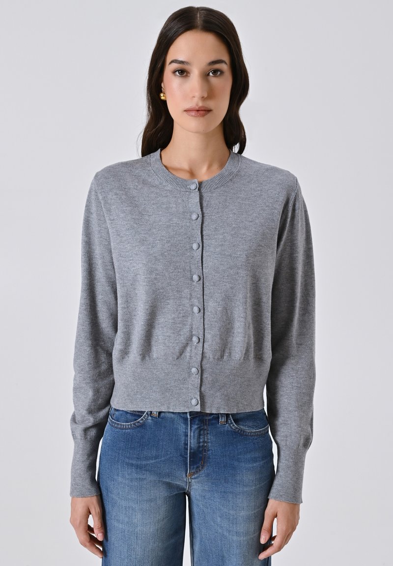 Giovane donna che indossa un cardigan grigio con bottoni e jeans blu, in piedi contro uno sfondo chiaro e neutro.