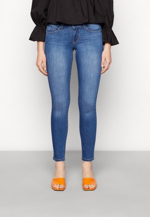 Frau trägt blaue Skinny-Jeans, schwarzen Langarmtop und orangefarbene offene Zehenabsätze und steht auf einem grauen Boden.